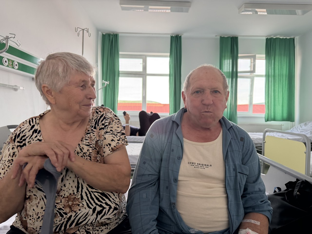 Two elderly patients sitting in a hospital room with medical equipment, illustrating healthcare needs for people over fifty requiring insurance coverage.