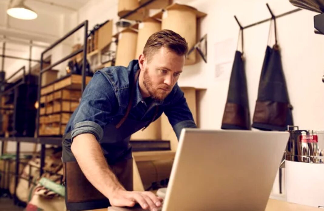 Small business owner at work exemplifies lean startup approach to building profitable directory ventures Man working on laptop at retail counter with shelving and hanging aprons behind him, representing the entrepreneurial approach to building scalable directory businesses with minimal resources.