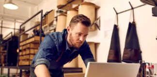Build a $1M Directory with Just $100 Man working on laptop at retail counter with shelving and hanging aprons behind him, representing the entrepreneurial approach to building scalable directory businesses with minimal resources.