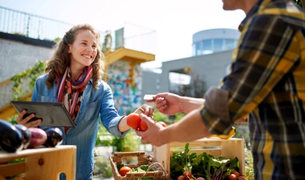 Direct customer engagement at farmers markets illustrates local commerce alternatives for neighborhood businesses Vendor accepts payment card from customer at outdoor farmers market with fresh produce in wooden crates, demonstrating direct engagement that builds neighborhood business connections.