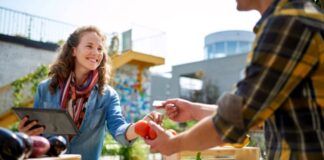 Why Your Neighbors Aren’t Buying From You Vendor accepts payment card from customer at outdoor farmers market with fresh produce in wooden crates, demonstrating direct engagement that builds neighborhood business connections.