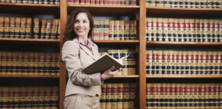 Law Business Directories vs Google: SEO Benefits Female attorney in beige suit holds an open book in a law library with floor-to-ceiling shelves, illustrating legal professional visibility and directory marketing.
