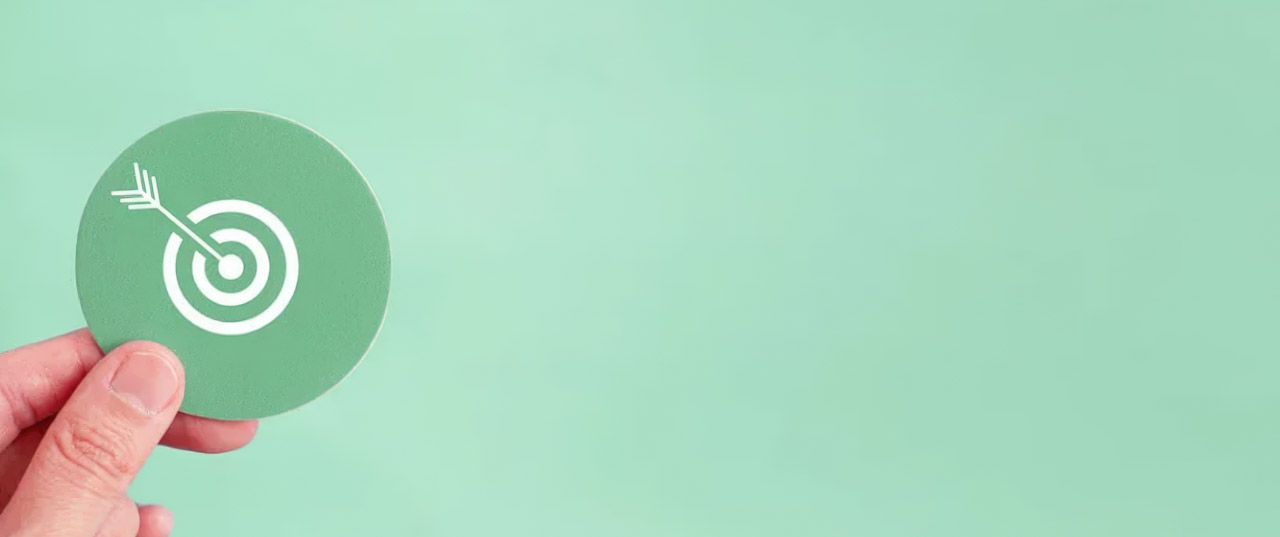 A hand holds a circular green card featuring a white target and arrow icon, symbolizing the precision required in identifying your target audience.