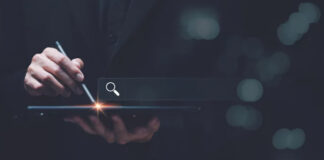 Hand holding stylus over tablet showing search icon with glowing interface, representing visual search and camera-driven discovery technologies in modern retail.