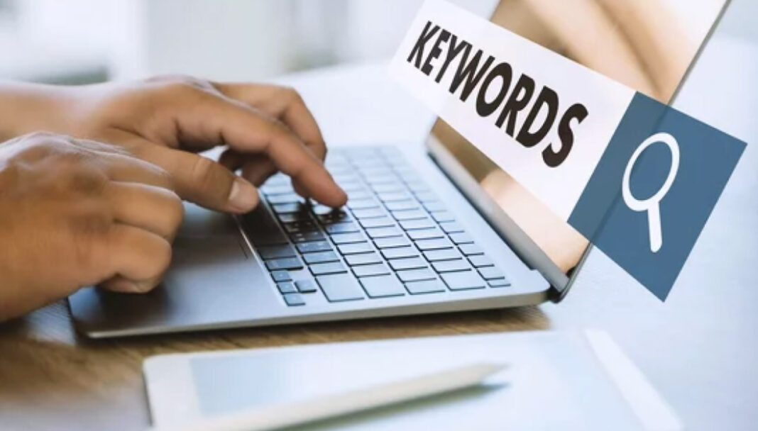 Laptop workspace illustrating keyword research and search methods for directory navigation Hands typing on a silver laptop keyboard with a keywords sign and magnifying glass icon visible, representing methods for finding and organizing directory search terms.