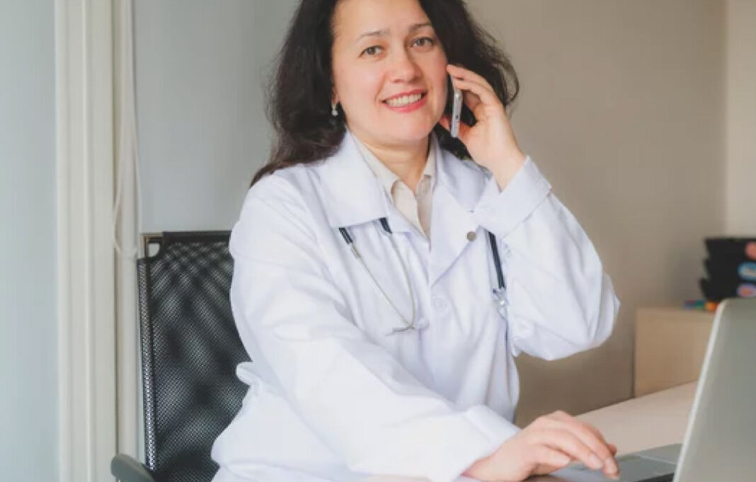 Medical professional uses smartphone, illustrating voice search accessibility for healthcare directory listings Healthcare professional in white lab coat smiles while speaking on smartphone at desk with laptop, representing voice search accessibility for medical directory listings.