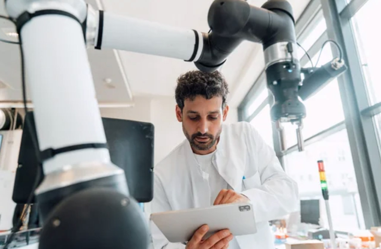 Laboratory technician holding tablet beside collaborative robotic arm in bright modern workspace, representing human-machine collaboration in content creation.