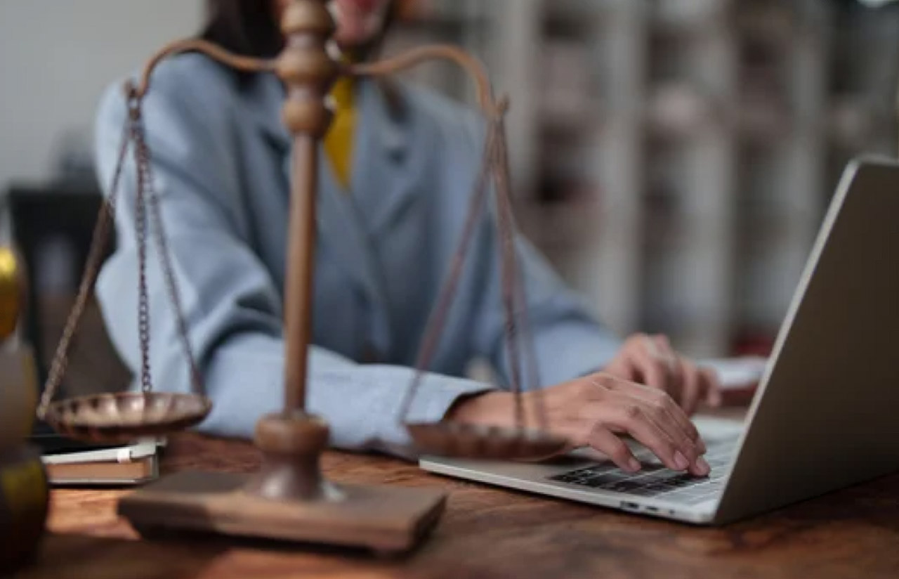 A legal professional types on a laptop at a wooden desk with bronze scales of justice in the foreground, illustrating digital visibility strategies for law firm directories.