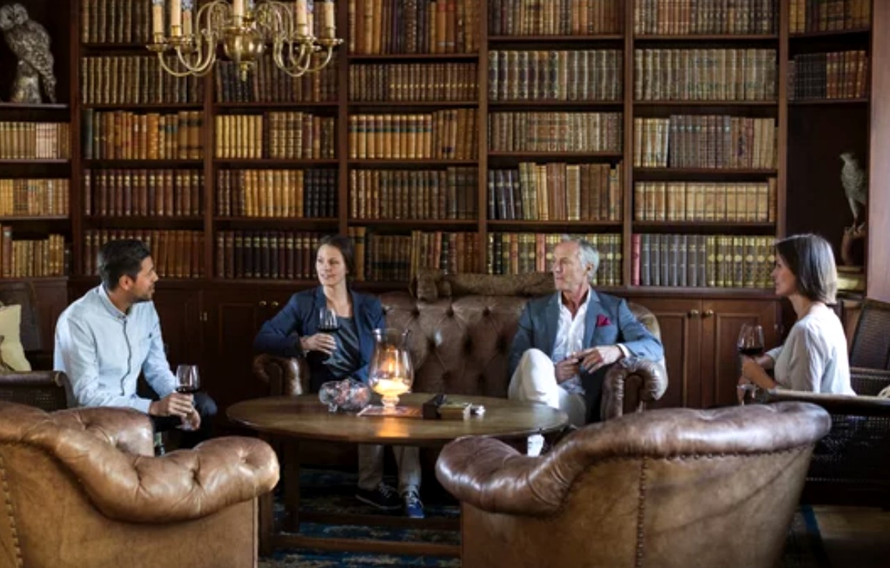 Four guests gather around a table in a traditional library with floor-to-ceiling bookshelves, illustrating the welcoming atmosphere that helps travel businesses attract direct bookings.