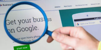 Why “Brand Lift” is Replacing “Click-Through” as the Primary KPI Hand holding magnifying glass examining Google business promotion on computer screen, symbolizing the detailed analysis of brand measurement replacing traditional click-through metrics.