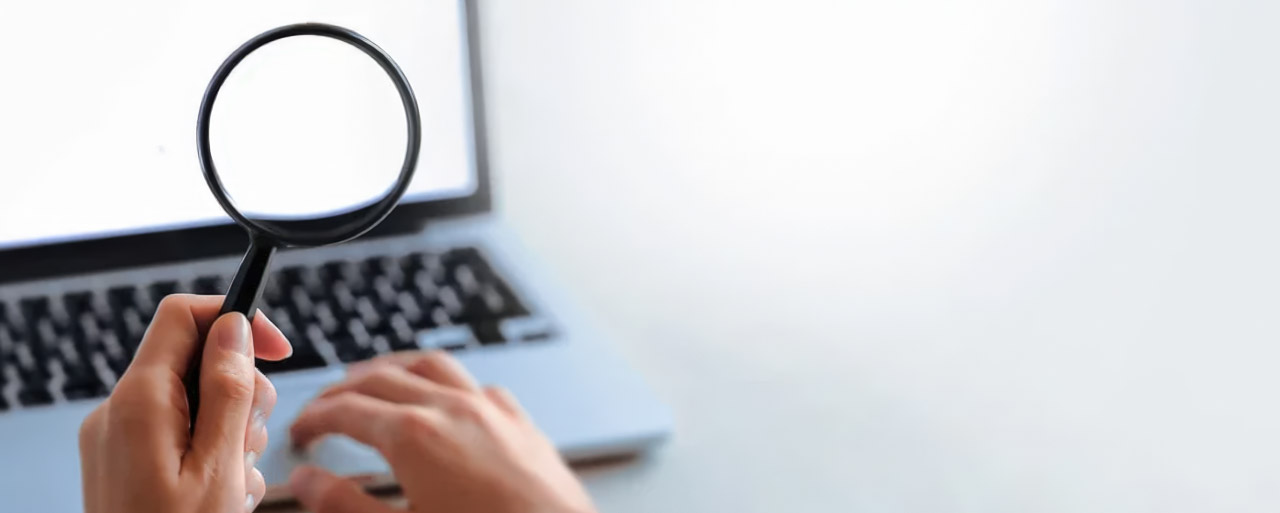 Person holding magnifying glass over laptop keyboard on white desk, symbolizing search and analysis in AI-powered SEO keyword research.