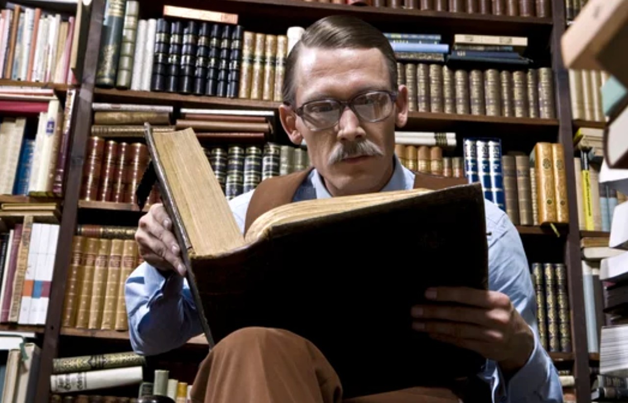 Man wearing glasses reads a large aged book surrounded by library bookshelves, symbolizing the research and knowledge that informs successful ebook creation and sales.