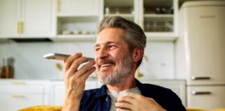 Man speaks into smartphone using voice assistant while seated on yellow couch in modern kitchen, illustrating voice search technology for content optimization.
