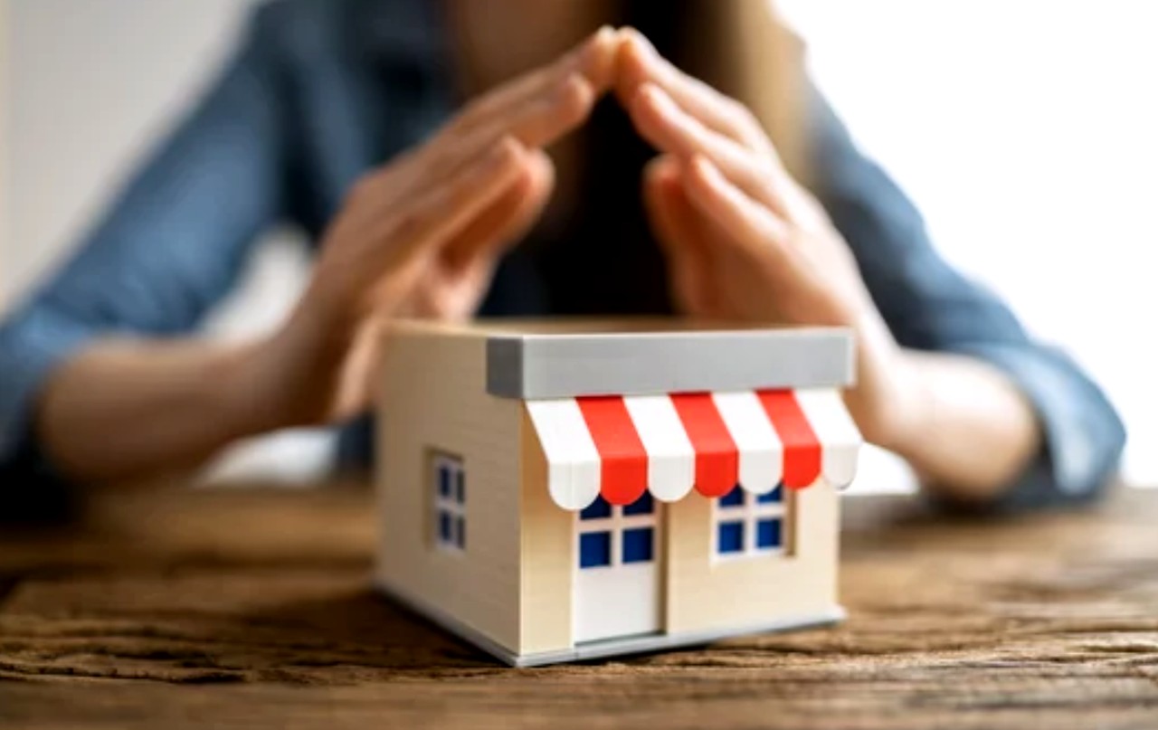 Hands hover protectively over a miniature shop model with red-and-white striped awning, symbolizing small business insurance protection and risk management.