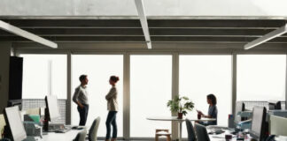 Modern open-plan office with professionals at workstations and in conversation near large windows, representing the professional credibility that small businesses establish through trusted presence.