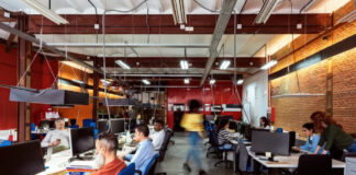 Open-plan office with employees working at computer workstations under industrial ceiling, representing the professional environment where businesses conduct directory research and competitive analysis.