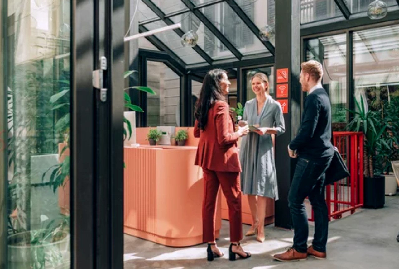Three professionals in business attire converse at a modern reception desk beneath a geometric glass ceiling, illustrating contemporary hospitality service environments.