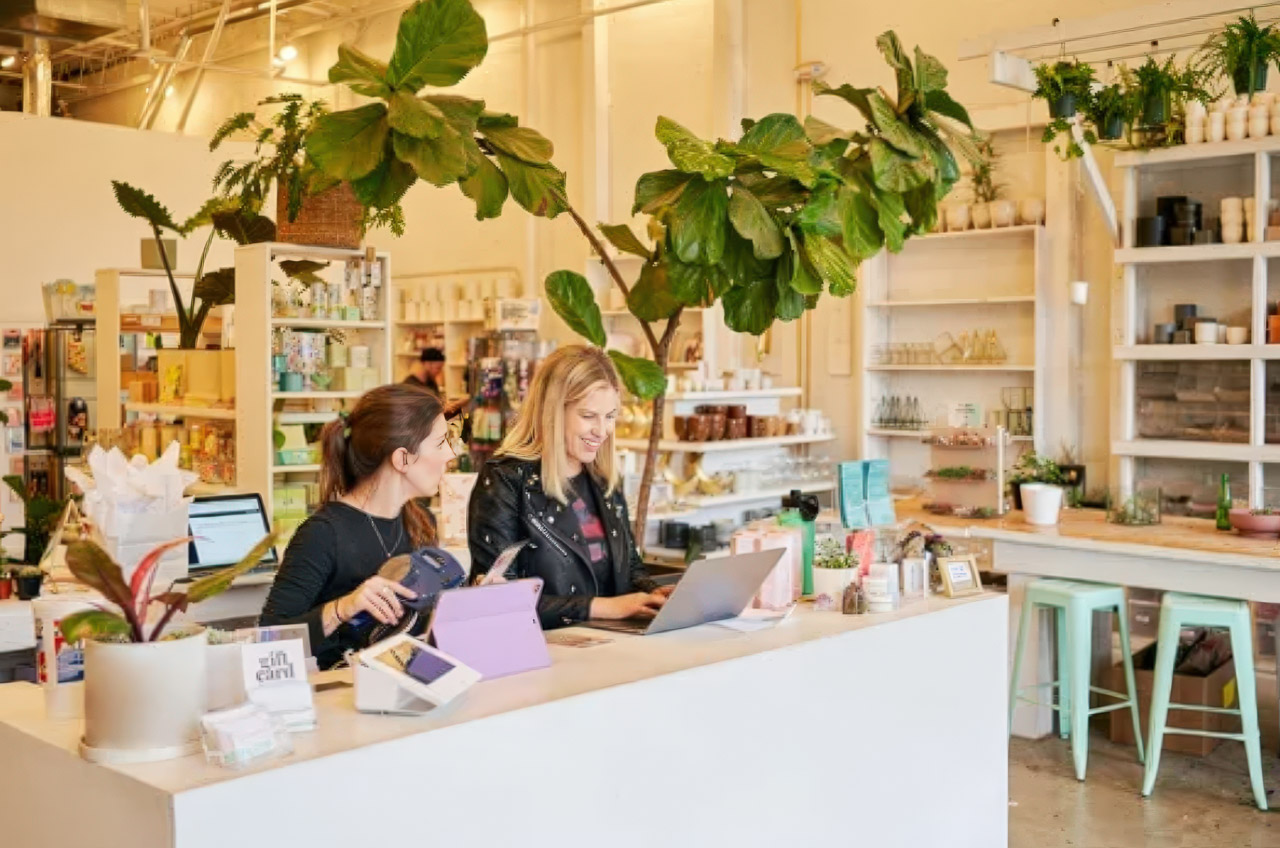 Two women work at a checkout counter in a bright modern boutique with plants and product displays, illustrating small business success through effective customer engagement and visibility.