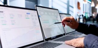 Managing Multiple US Directories Made Simple Three laptops on an office desk display business scheduling software while a hand with pen points at the center screen, illustrating multi-platform directory coordination.