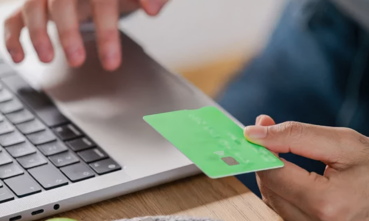 Hands hold a green payment card near a laptop keyboard on wooden desk, illustrating digital commerce and online business transactions.