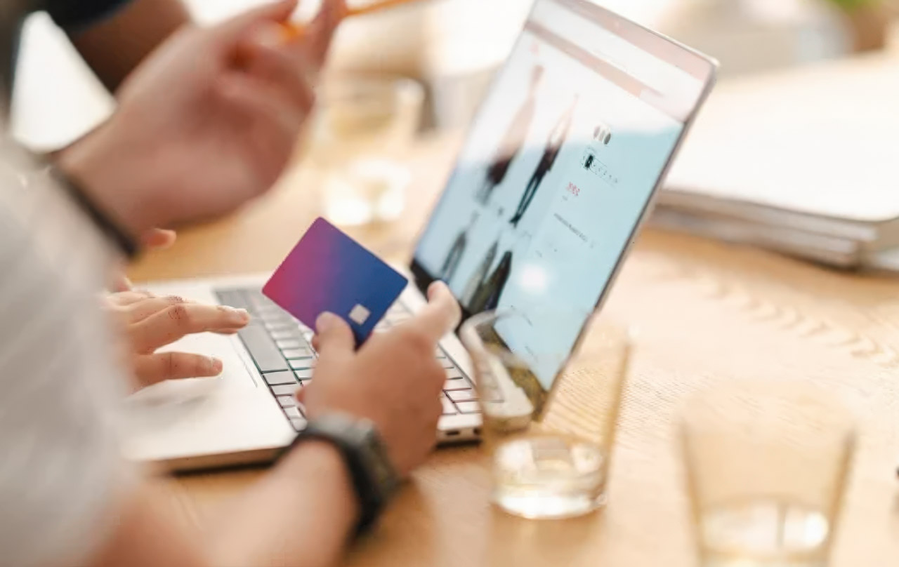 Person holding purple credit card while shopping online with laptop at wooden desk, representing modern consumer search and purchase behavior that businesses capture through directory listings.