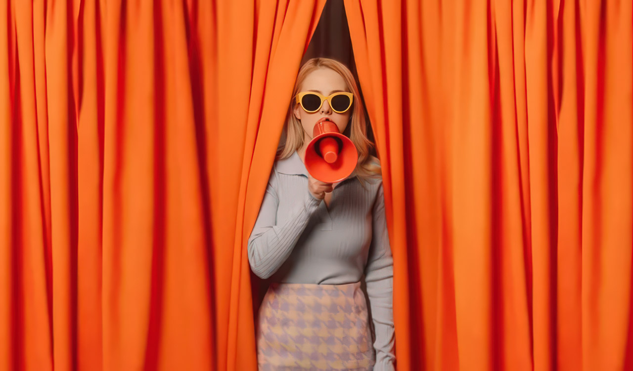 Person holds red megaphone between orange curtains in studio portrait, illustrating the amplification of brand messages through impression-based marketing strategies.