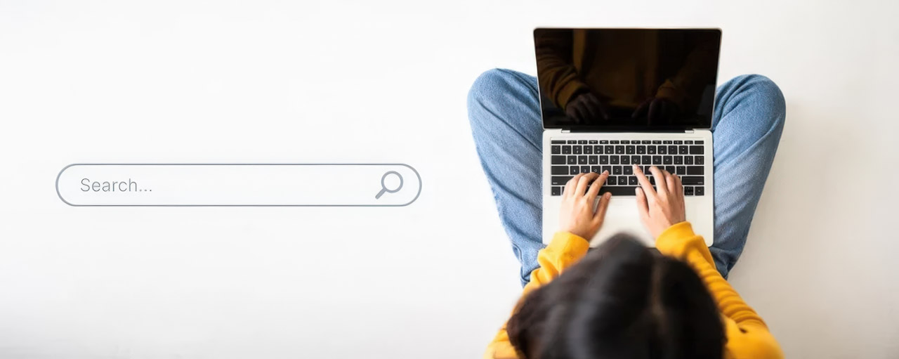 Overhead view of person in yellow sweatshirt typing on laptop with search bar graphic overlay, illustrating search engine optimization research and digital marketing work.
