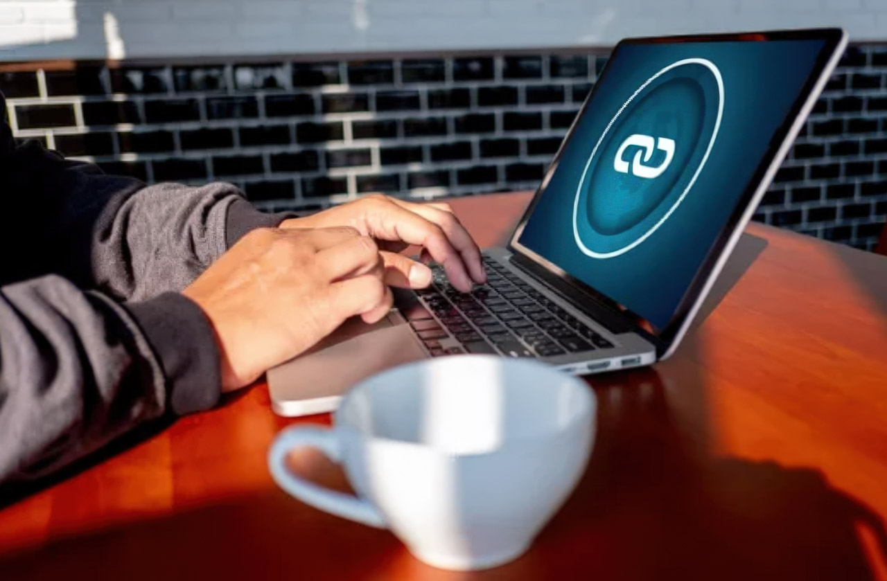 Person typing on silver laptop with branded screen at wooden table with coffee cup, illustrating the focused work required for website troubleshooting and performance optimization.