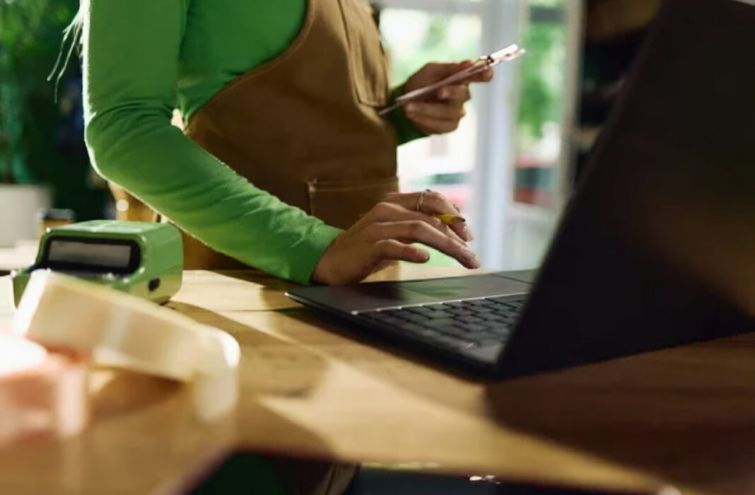 Multi-device workspace illustrating the importance of mobile-first indexing for modern business directories Person simultaneously using smartphone and laptop at wooden workspace with point-of-sale device, demonstrating the multi-device approach central to mobile-first indexing strategies.