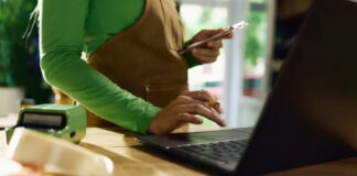 Person simultaneously using smartphone and laptop at wooden workspace with point-of-sale device, demonstrating the multi-device approach central to mobile-first indexing strategies.