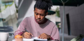 The Easiest Way to Get Listed Person writes in notebook at table with coffee and phone, representing the process of gathering information for business directory listings.