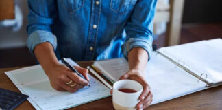Submit to 100 Directories in 60 Minutes (Step-by-Step) Person in denim shirt writes in notebook at wooden desk with calculator, papers, and beverage, representing organized workspace for systematic directory submission tasks.