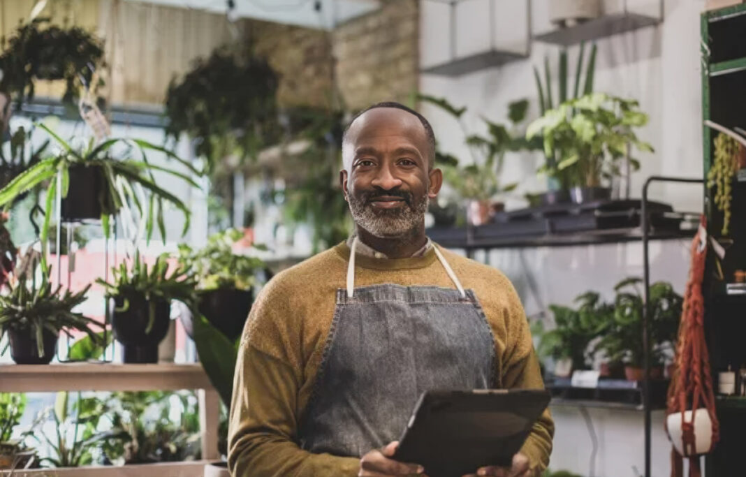 plant-nursery-owner-tablet-digital-commerce-sustainable-retail Plant shop owner in apron holds tablet among potted plants in nursery, representing sustainable business practices and transparent communication with customers.