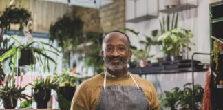 Plant shop owner in apron holds tablet among potted plants in nursery, representing sustainable business practices and transparent communication with customers.