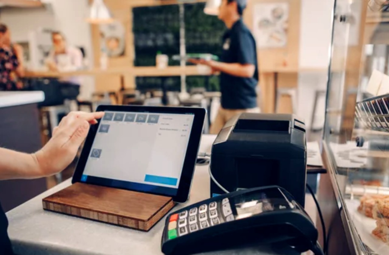 A hand operates a tablet point-of-sale system with credit card terminal and receipt printer on a café counter, representing payment-enabled business directory infrastructure.