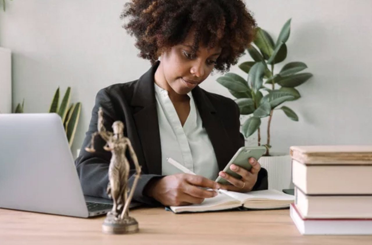 Professional woman at wooden desk writing in notebook while holding smartphone, with laptop and organized workspace visible, representing entrepreneurs managing business directory listings.