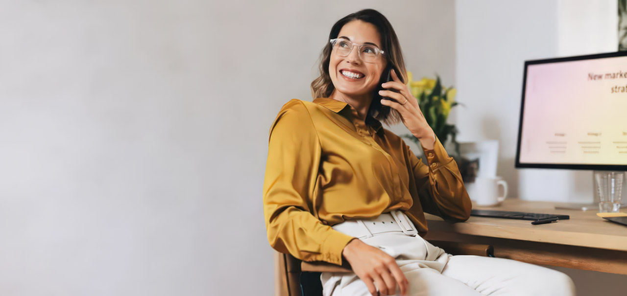 Smiling professional woman in eyeglasses takes a phone call at her office desk with computer monitor, representing modern predictive customer service operations.