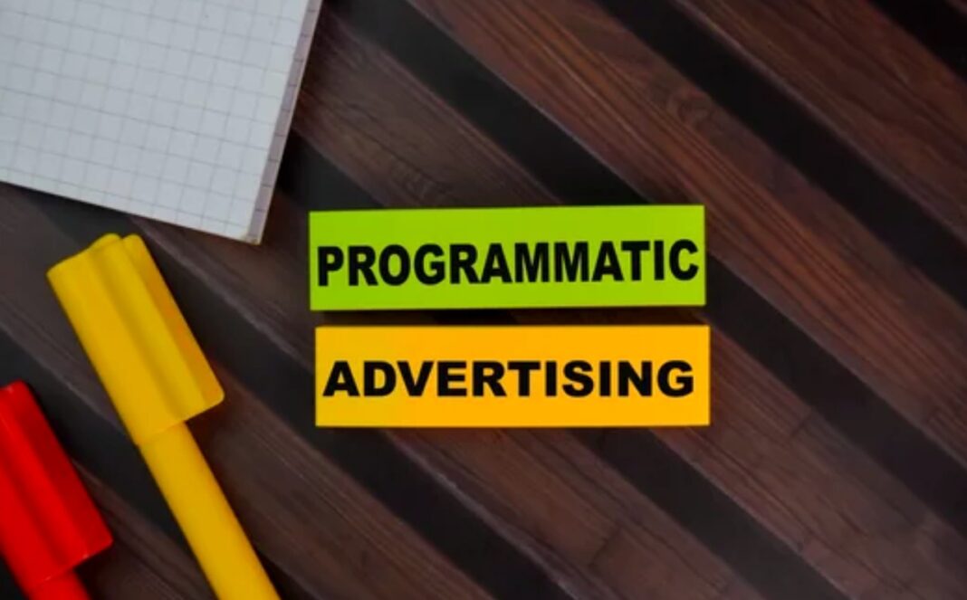 Programmatic advertising concept illustrates automated native advertising evolution in the AI era Flat lay photograph of a programmatic advertising text banner on a wood surface with colored blocks and graph paper, representing automated advertising strategies in digital marketing.