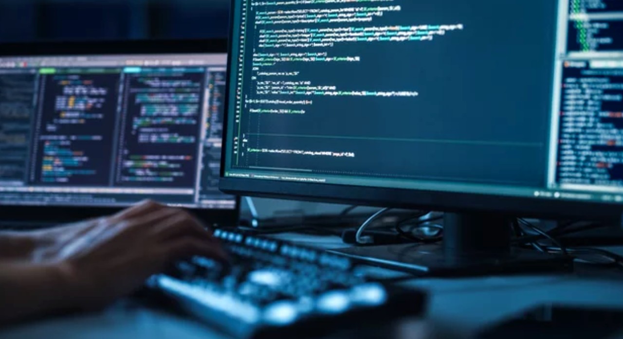 Programmer's hand on backlit keyboard with multiple monitors displaying code in cyan text on dark backgrounds, illustrating workspace for Python SEO automation.