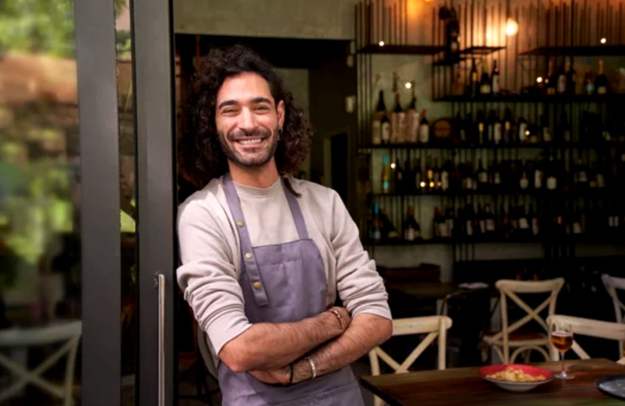 Restaurant staff member in gray apron stands at entrance with crossed arms and welcoming smile, representing successful restaurant directory listing practices.