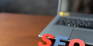 Three-dimensional red and blue letters spelling SEO stand on a wooden desk in front of a laptop, representing search engine optimization strategies and ranking factors.