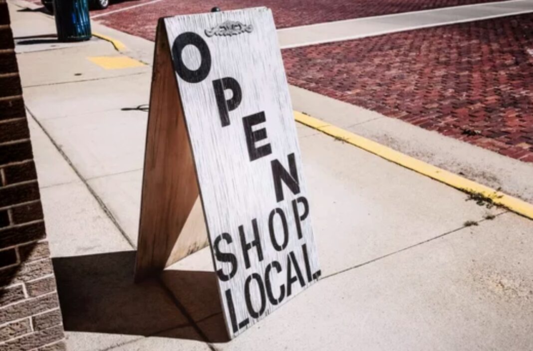 Sidewalk signage invites customers to shop local, illustrating grassroots marketing for small businesses A wooden A-frame sign reading 'OPEN SHOP LOCAL' on a sidewalk beside a brick building, illustrating local business marketing strategies.