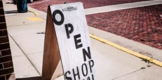 How do I market my local business? A wooden A-frame sign reading 'OPEN SHOP LOCAL' on a sidewalk beside a brick building, illustrating local business marketing strategies.