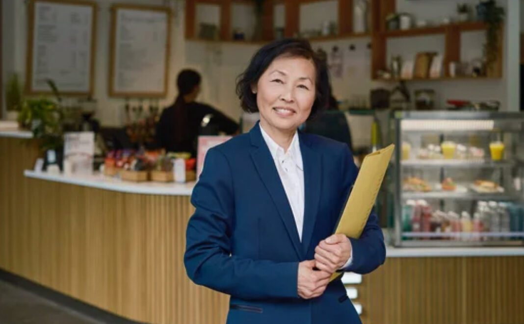 Local café owner represents the entrepreneurial spirit driving neighborhood commerce and business visibility Professional woman in business suit holding folder stands in café interior, representing local business ownership and entrepreneurial presence in neighborhood commerce.