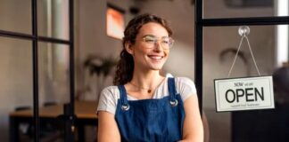 Woman wearing blue apron and glasses stands with crossed arms at glass storefront entrance with open sign, representing local business ownership and community retail.