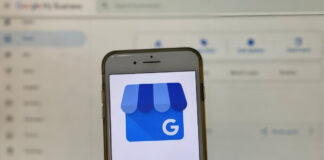 Smartphone displaying Google My Business app icon held in front of computer screen, demonstrating digital tools for managing NAP consistency and business directory listings.