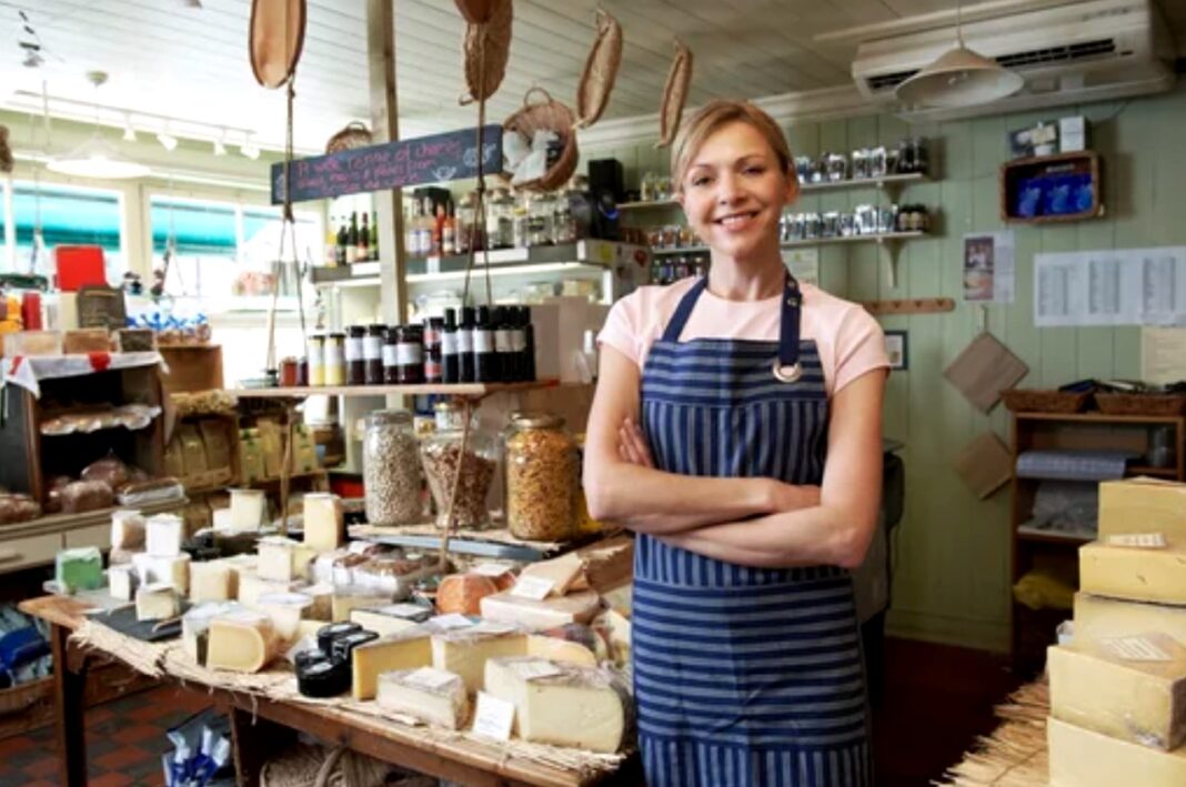 Independent food retailer exemplifies niche marketing through specialized product curation and personal service Woman in striped apron behind wooden cheese counter in specialty food shop, illustrating focused niche marketing and personalized customer service in independent retail.