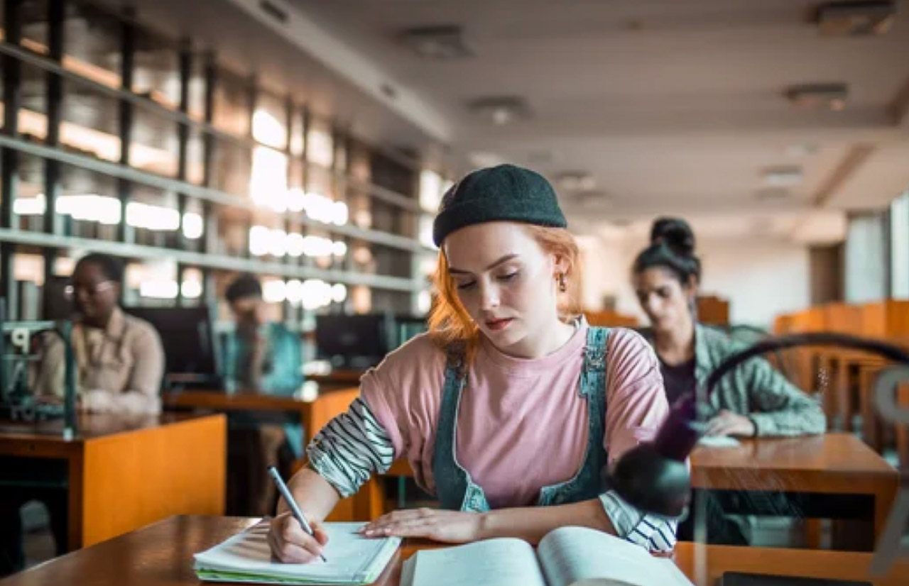 A student writes in a notebook at a library table surrounded by bookshelves, representing the human editing and revision process central to refining AI-generated content.