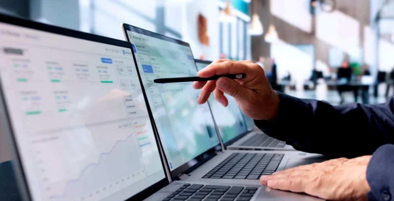 Hand holding stylus points at laptop screen showing business dashboard in bright office workspace, representing the hands-on technical audit process.