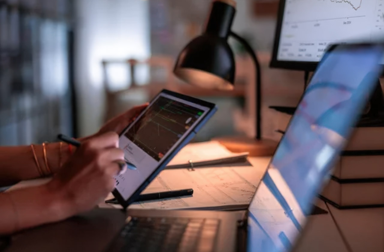 Hands using a tablet displaying graphs on a wooden desk with lamp and books, representing digital tools for navigating English-friendly Asian business directories.